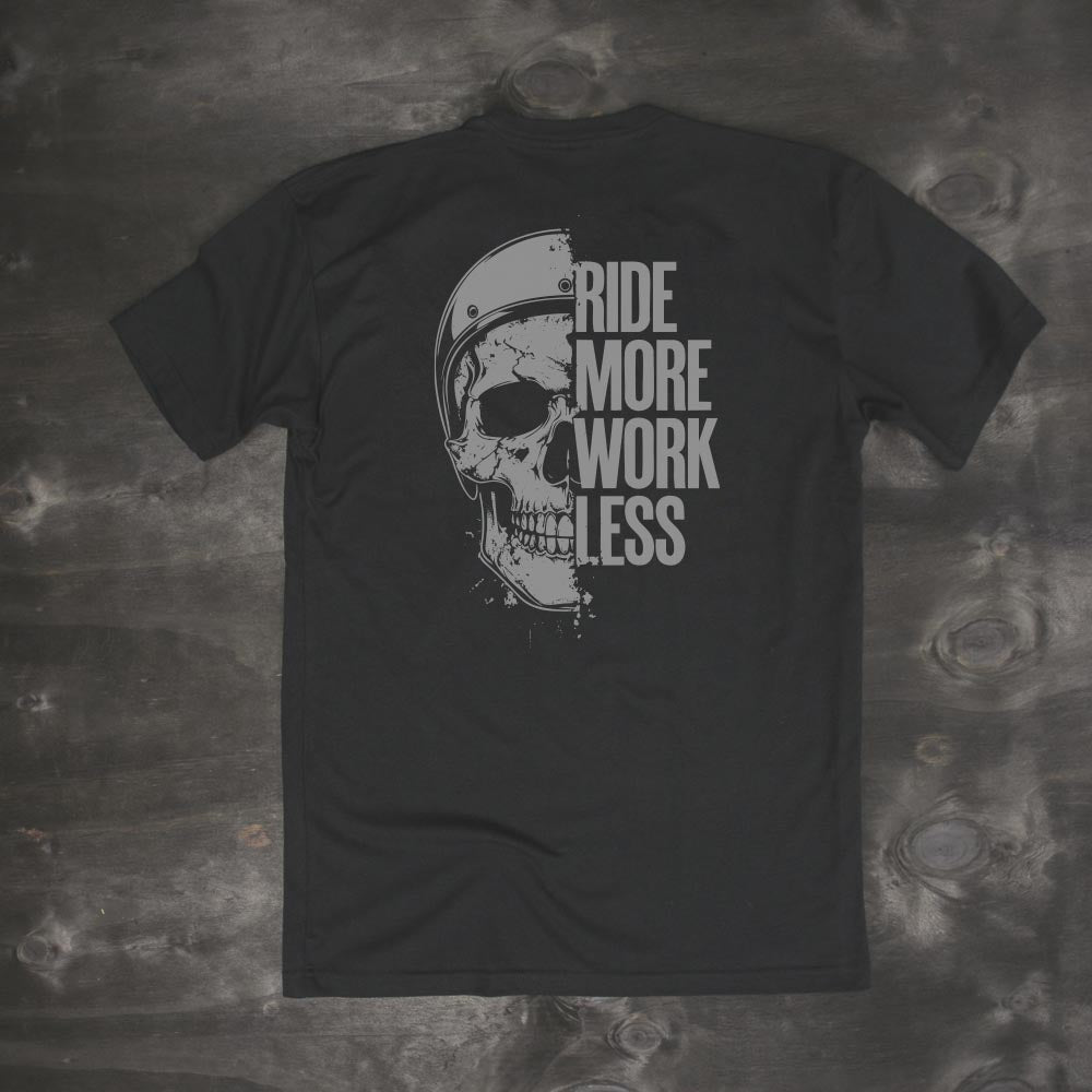 Men's black t-shirt with skull and helmet graphic and bold text 'Ride More Work Less' on the back, laid flat on dark wood background