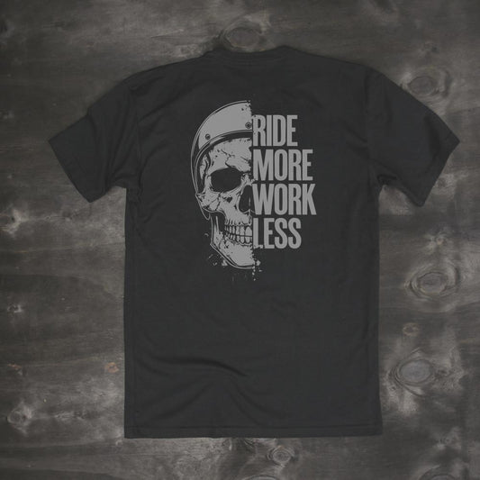 Men's black t-shirt with skull and helmet graphic and bold text 'Ride More Work Less' on the back, laid flat on dark wood background