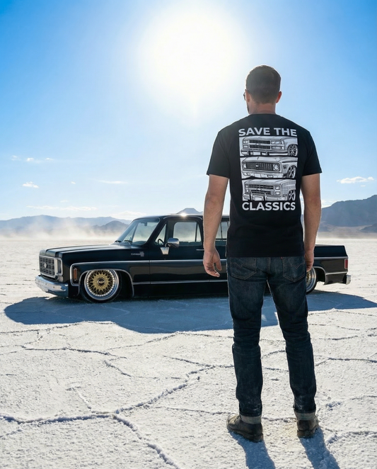 Man wearing a black t-shirt with a graphic design and text, standing in front of a classic car on a desert road.