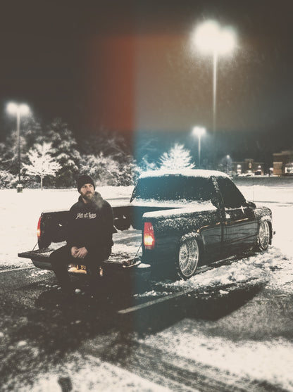 Person sitting next to a snow-covered truck in a snowy parking lot at night.